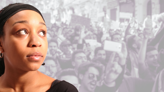 The background is a black and white semi-transparent image of many people involved in a protest, holding placards and shouting aggressively. Set against this, in full colour, is a Black female looking off into the distance and looking anxious and pensive, as if she is viewing the scene in her head.